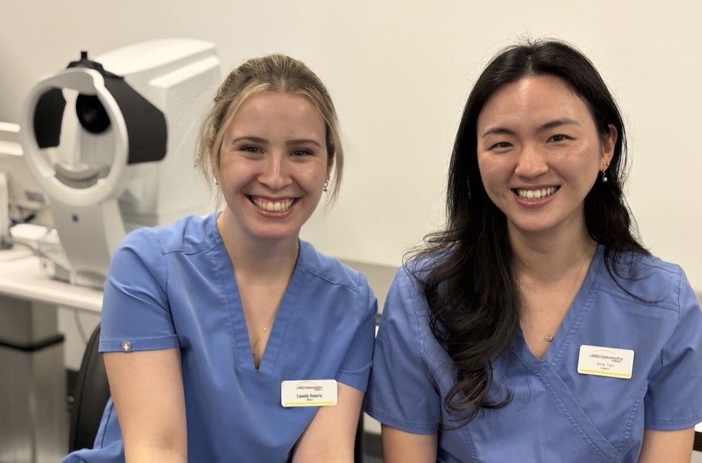 Two young women wearing blue scrubs and name tags are seated in a clinic with medical equipment visible in the background