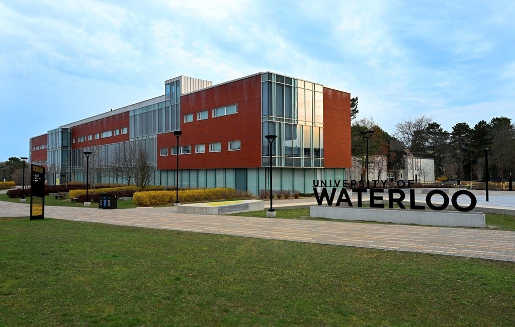 University of Waterloo signage in the foreground with Tatham Centre in the background on a summer cloud filled day