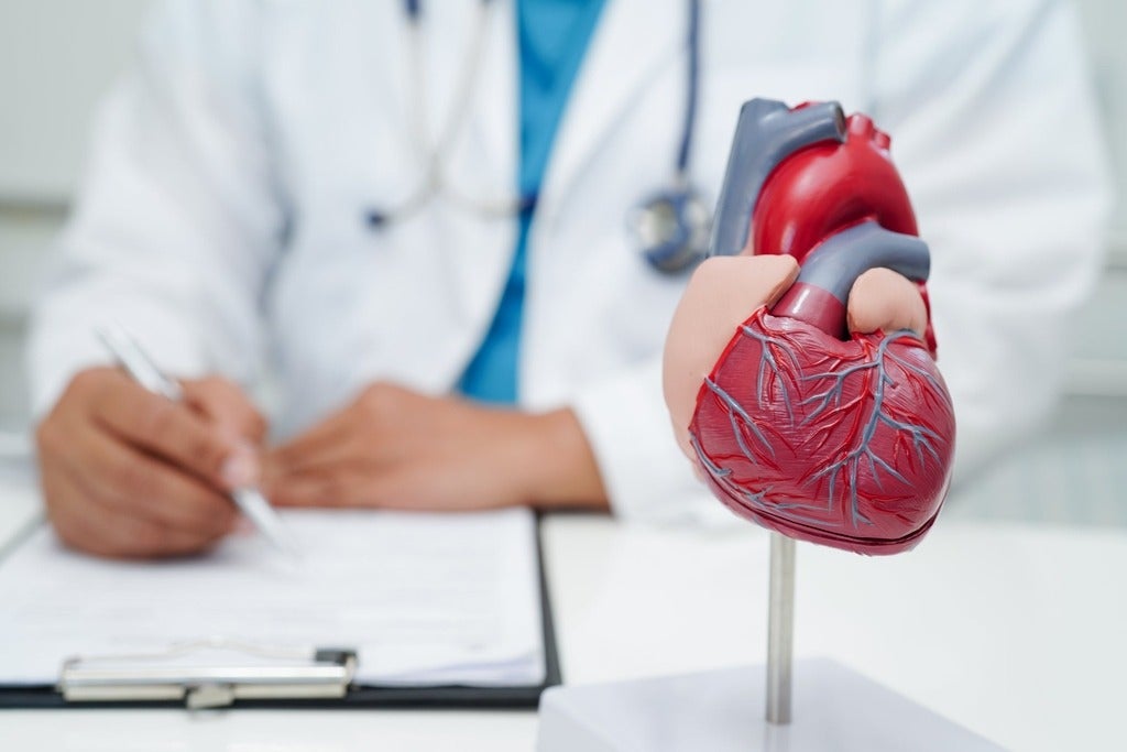 Doctor making notes on clipboard on desk with model of a human heart in foreground.