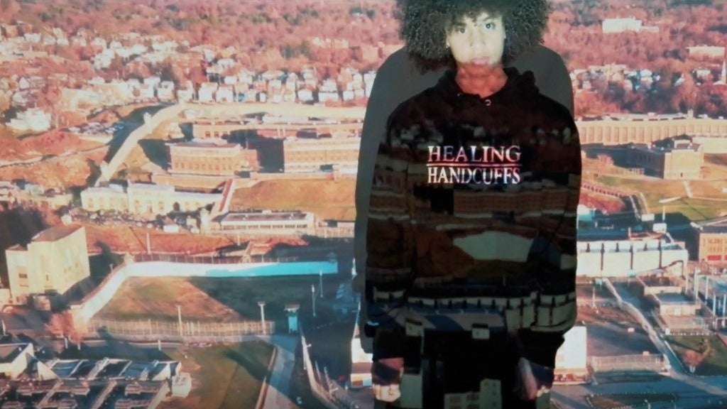 A Black teenager wearing a hoodie that says "Healing Handcuffs" stars at the camera, an image of a prison projected across him
