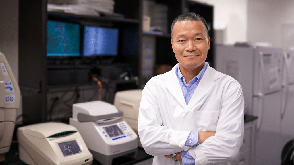 Juewen Liu is wearing a white lab coat and blue collared shirt. He is standing with his arms crossed and smiling in his lab.