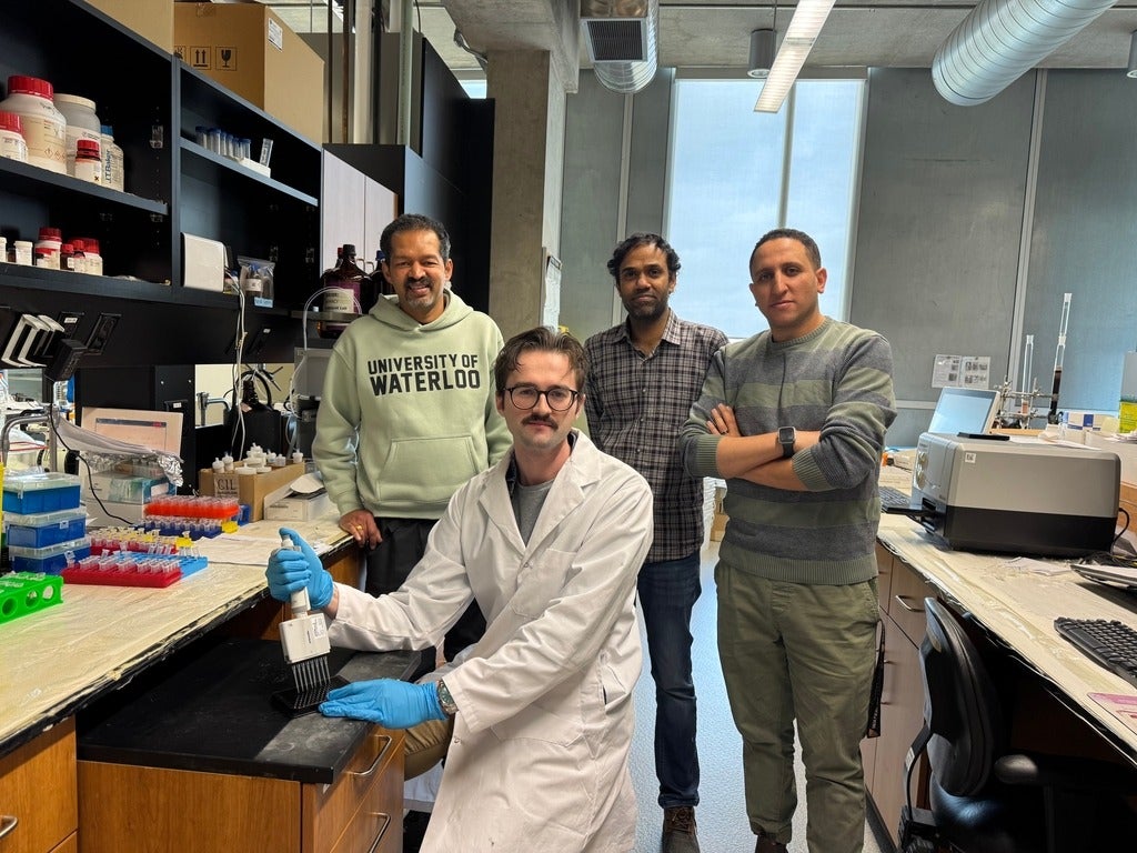 Four people in a lab, with one of them seated and wearing a lab coat and gloves
