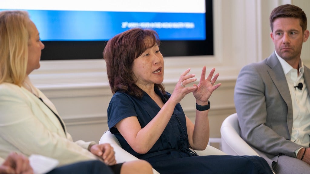 Jennifer Lee speaking with her hands in motion while two colleagues listen on each side and a big screen behind