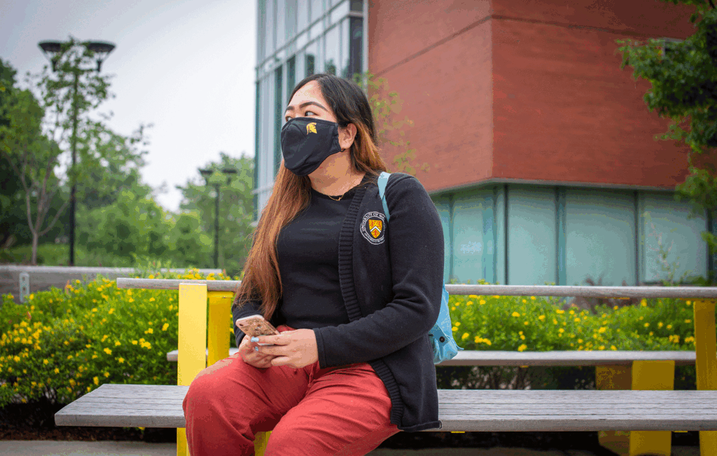A student sits on a park bench on campus with their mobile phone.