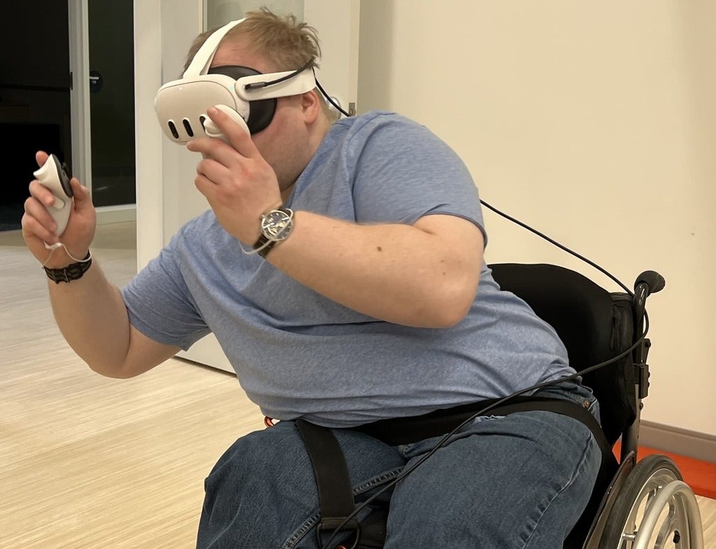 A person in a wheelchair wearing VR googles holding a controller leaning to avoid hazards in the VR game, Beat Saber.