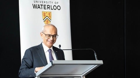 President Goel smiles as he reviews notes on a podium in front of a UWaterloo banner.