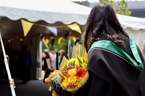 back of graduate wearing robes, green hood and carrying flowers
