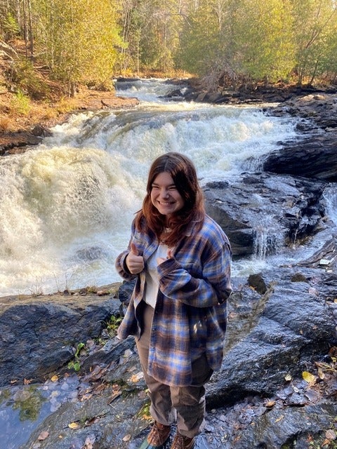 A smiling young woman wearing a  plaid shirt stands in front of a mountain stream