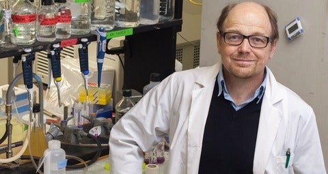 Professor wearing lab coat stands in front of a shelf of beakers in a laboratory