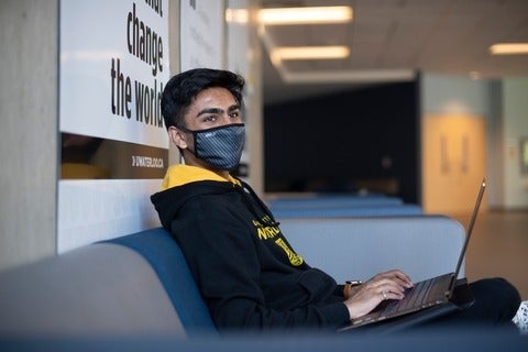 A student sitting in a Waterloo hoodie and mask while typing on a laptop