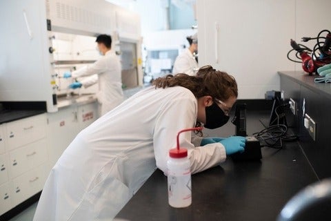 Researcher in lab looks through microscope while others work in the lab