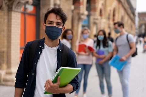 A student wearing a face mask and holding a book with other students in the background