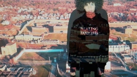 A Black teenager wearing a hoodie that says "Healing Handcuffs" stars at the camera, an image of a prison projected across him