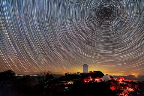 A long-exposure image captures circular star trails above telescope domes illuminated in red light on a dark mountain.