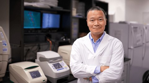 Juewen Liu is wearing a white lab coat and blue collared shirt. He is standing with his arms crossed and smiling in his lab.