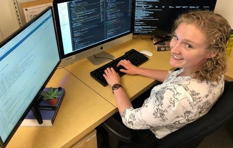 Pascale Walters smiles up at the camera as she sits in front of computer screens with code