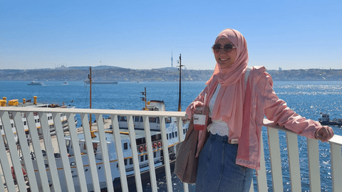Sara is leaning against a white fence with the ocean in the background.