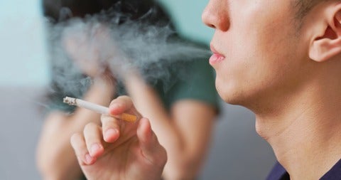 Man exhales cigarette smoke while woman sits in background