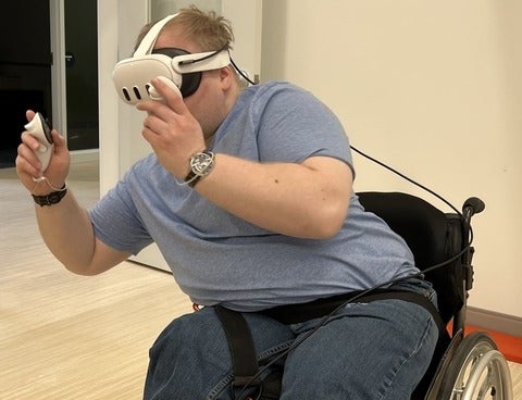 A person in a wheelchair wearing VR googles holding a controller leaning to avoid hazards in the VR game, Beat Saber.