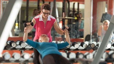Older adult lifts weights in gym while supervised by personal trainer.