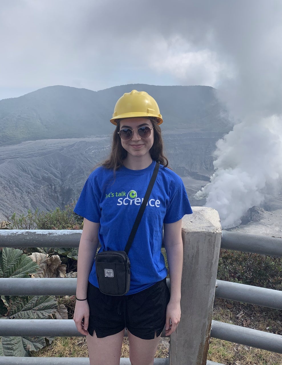 Sydney standing in front of an volcano site. She is wearing a yellow contruction helmet and a blue t-shirt that says "Let's talk science". 