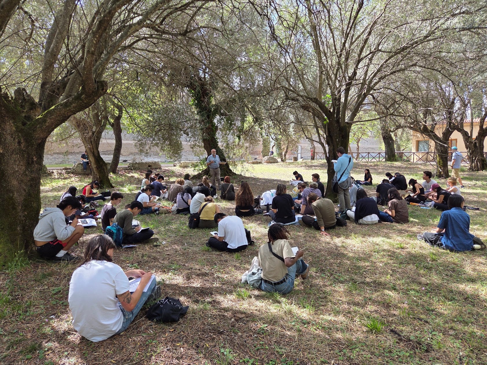 School of Architecture cohort in Rome sitting on the ground of a park, sketching on notepads while listening to Rick Haldenby talk 