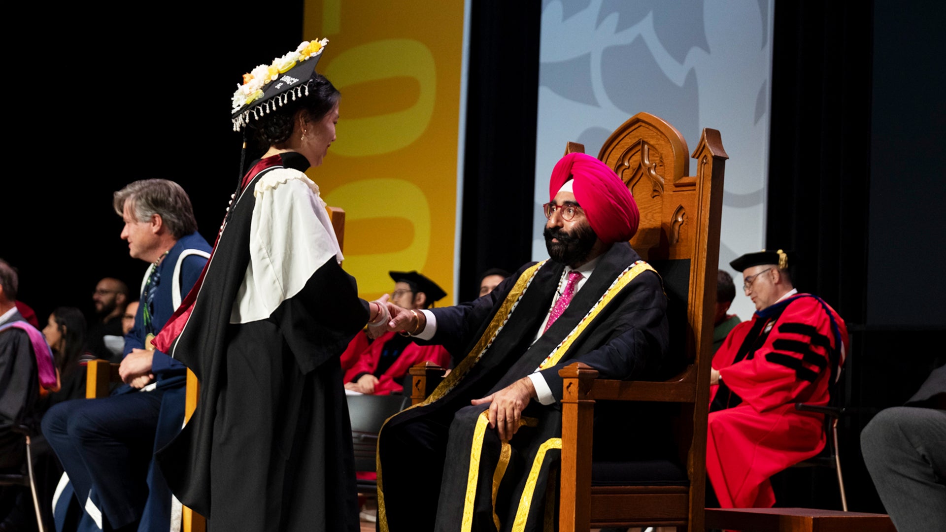 Chancellor Bachher shakes hands with graduate in elaborated adorned regalia 