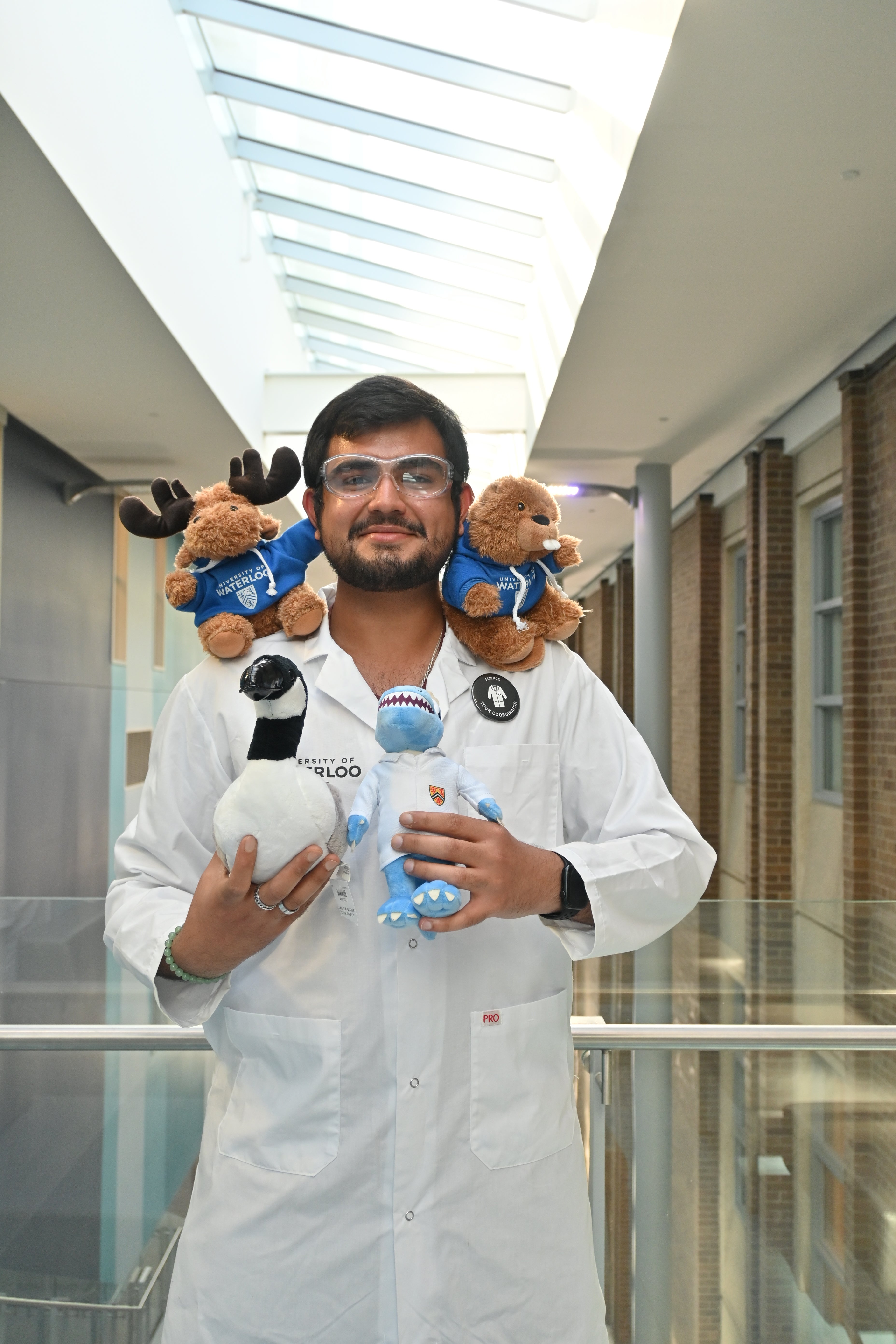 Ayush Verma wearing a labcoat and posing with four plushies including Waterloo Mr. Goose, Cobalt the mascot, Ren the Moose and beaver in a Waterloo Science hoodie 
