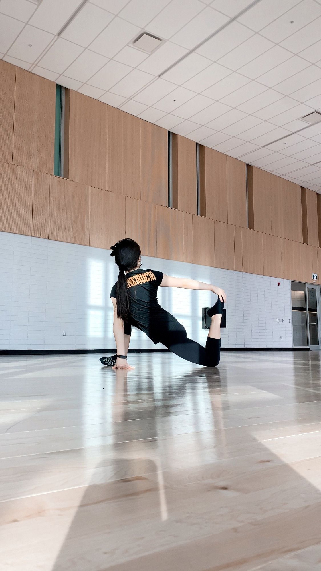 Cindy Wei in a kneeling lunge stretch in a fitness studio. She is facing the back of the room and has 