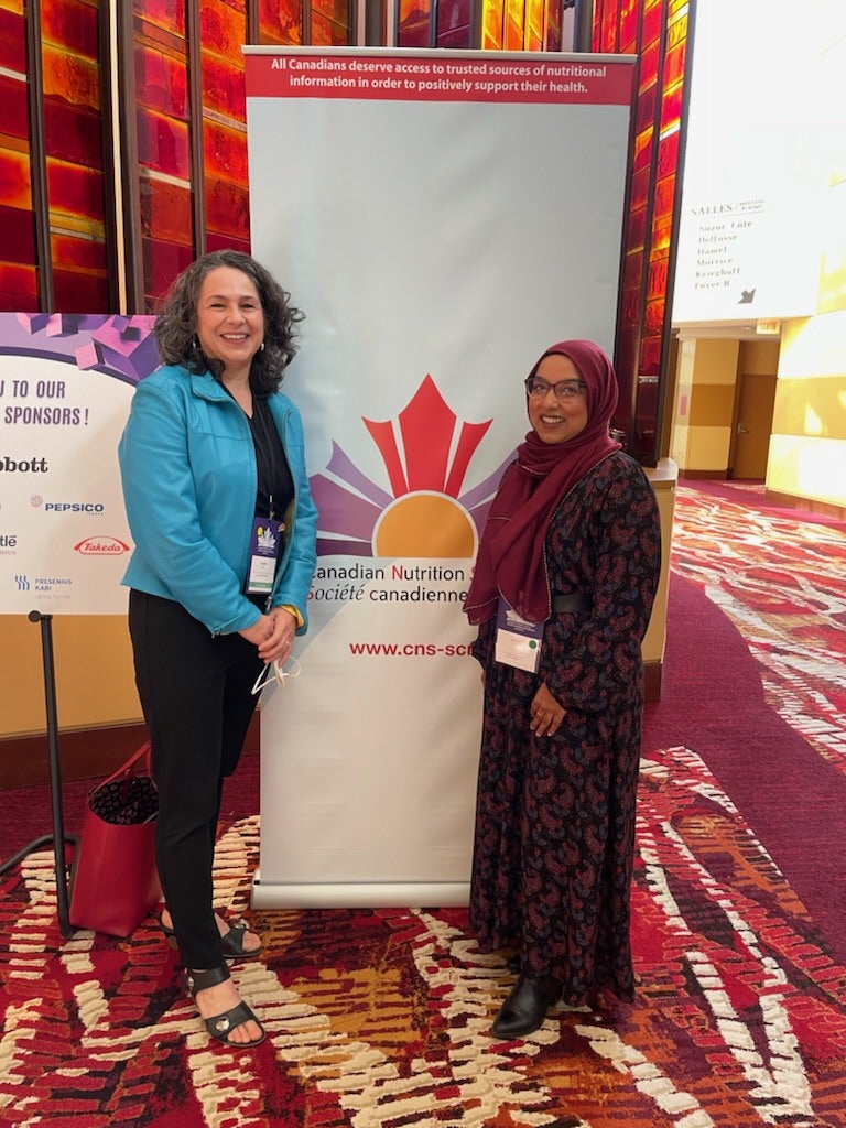 Heather Keller and Safura Syed standing in front of Canadian Nutrition Society conference banner