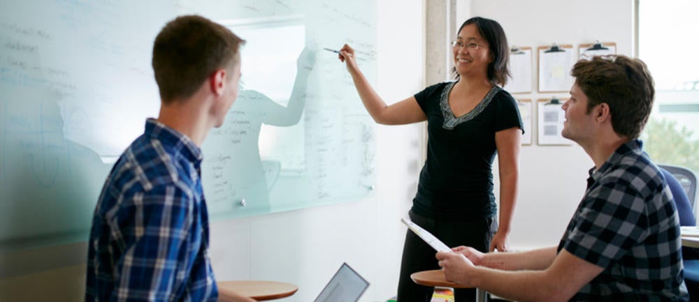 Professor at whiteboard speaks to two students.