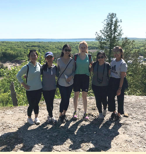 Five young women stand on a rocky outcrop overlooking a lake