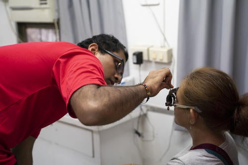 A man in a red shirt conducts trial frame refraction while examining a woman’s eyes