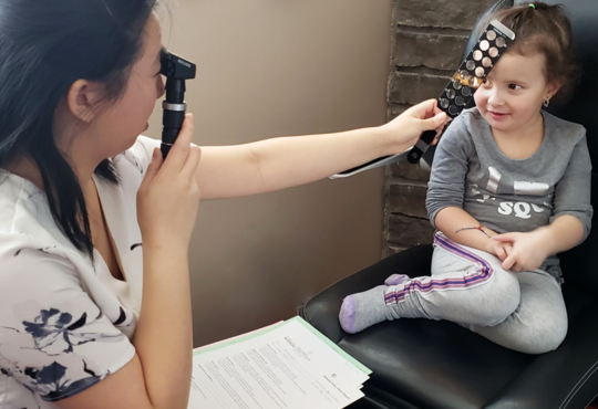 Rachel Ng looks through a retinoscope while holding a lens bar to the eye of a small girl.  