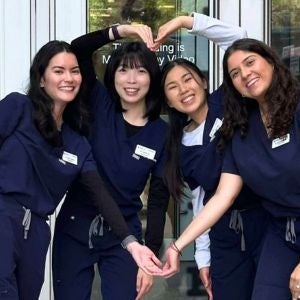 Four young women in scrubs form a heart with their arms