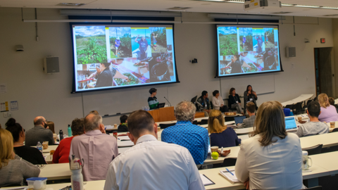 Five presenters sitting below two large screens listen to a speaker