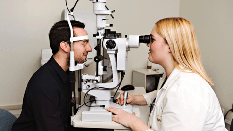 A young blond woman in a clinic coat examines a man’s eyes using a slit lamp.