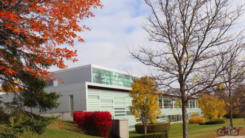 University of Waterloo School of Optometry and Vision Science surrounded by vibrant fall foliage, with bright red and orange leaves on trees and shrubs in the foreground, yellow trees along the lawn, and a bicycle parked beside a sidewalk under a partly cloudy sky.