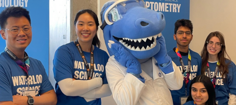A group of Waterloo Science student ambassadors stands with a large blue shark mascot wearing safety goggles and a white lab coat. The ambassadors wear blue Waterloo Science shirts and event lanyards, posing in front of School of Optometry and Vision Science display banners.