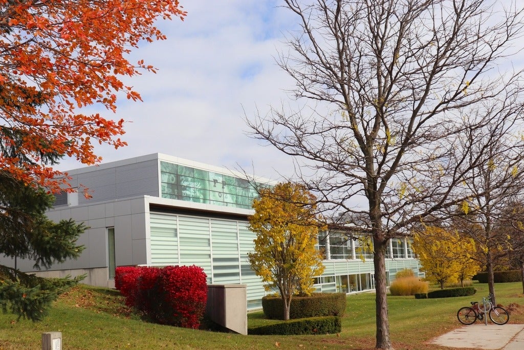 University of Waterloo School of Optometry and Vision Science surrounded by vibrant fall foliage, with bright red and orange leaves on trees and shrubs in the foreground, yellow trees along the lawn, and a bicycle parked beside a sidewalk under a partly cloudy sky.