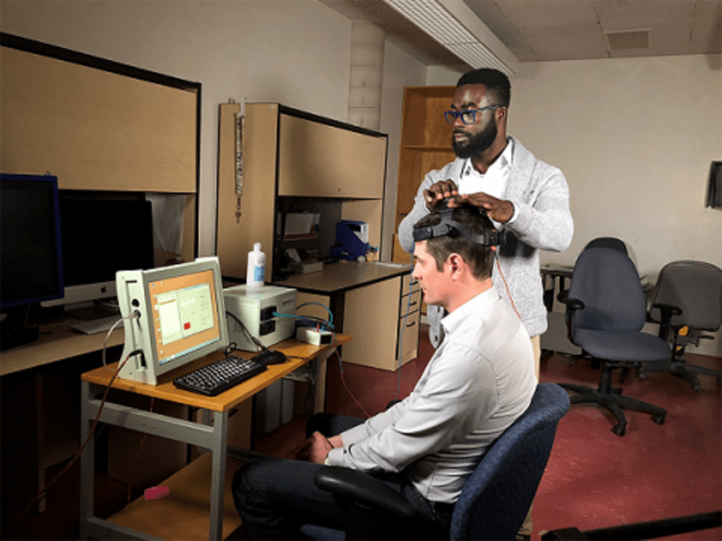 Richard Donkor looks at a computer monitor while adjusting Dr. Ben Thompson’s headset