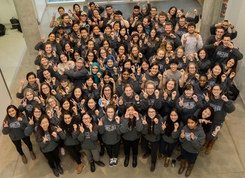 A crowd of Optometry students wearing grey hoodies wave at the camera above them