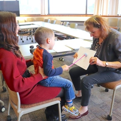 Child on mother's lap, pointing with pointer at paper held by optometrist