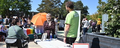 A PACS student speaks with a PACS professor, who wears a green t-shirt that has "designated dialoguer" written on the back during a community event in the Arts Quad on a warm fall day.