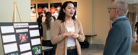 A PACS student speaks with a research fellow during a research exhibit in the Grebel atrium. The student explains her research project titled "Cafe Femenino", on a nearby display board.