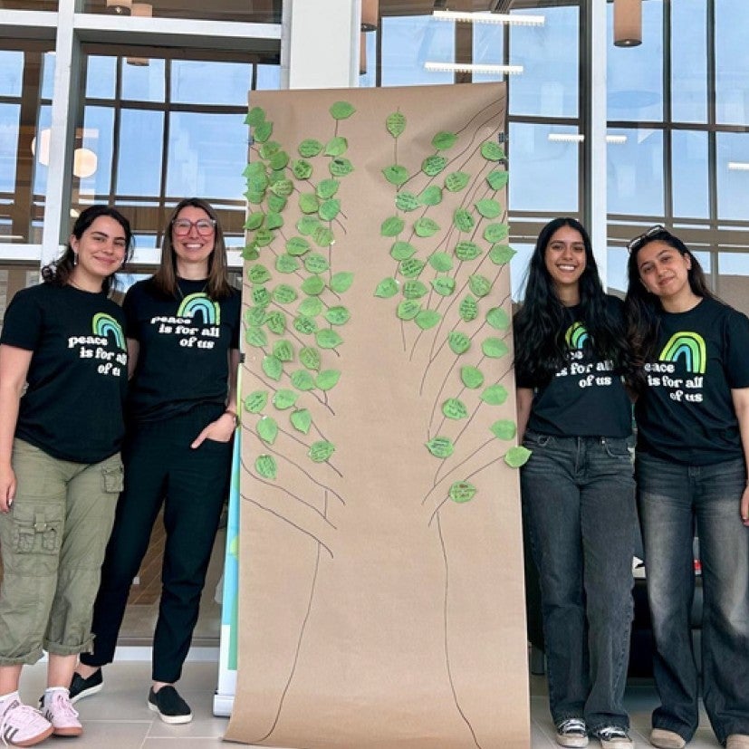 Zelal, Sarah, Zayna and Aala pose by a big drawn tree that people have added their own leaves and comments to