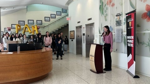 A person stands at a podium with a microphone in a bright building lobby decorated with large floral wall art. A University of Waterloo banner reading “Innovating Pharmacy Together” is positioned beside the podium. In the background, a group of people stands near a staircase, facing the speaker. Gold balloon letters forming “PAM” and two heart-shaped balloons are displayed behind the group near the reception desk. Two elevators and wall-mounted plaques are visible along the back wall.