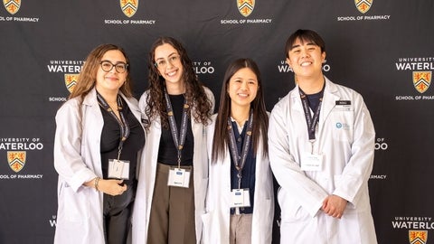 Four people wearing white lab coats stand smiling in front of a University of Waterloo School of Pharmacy backdrop, each wearing a conference name badge.