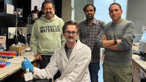 Four people are positioned in a laboratory setting with benches, instruments, and lab supplies visible. One person in the foreground sits at a lab bench wearing a white lab coat and blue gloves while holding a pipette over laboratory equipment; three others stand behind with arms relaxed or crossed. Shelves, bottles, tubes, and devices surround the workspace, and a sweatshirt in the background displays the text “University of Waterloo.”