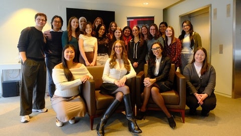 A group of about twenty-five people pose together indoors in a bright, modern room with beige walls. Three people sit on armchairs at the front, while the rest stand closely behind them. An elevator door and a standing banner are visible in the background.